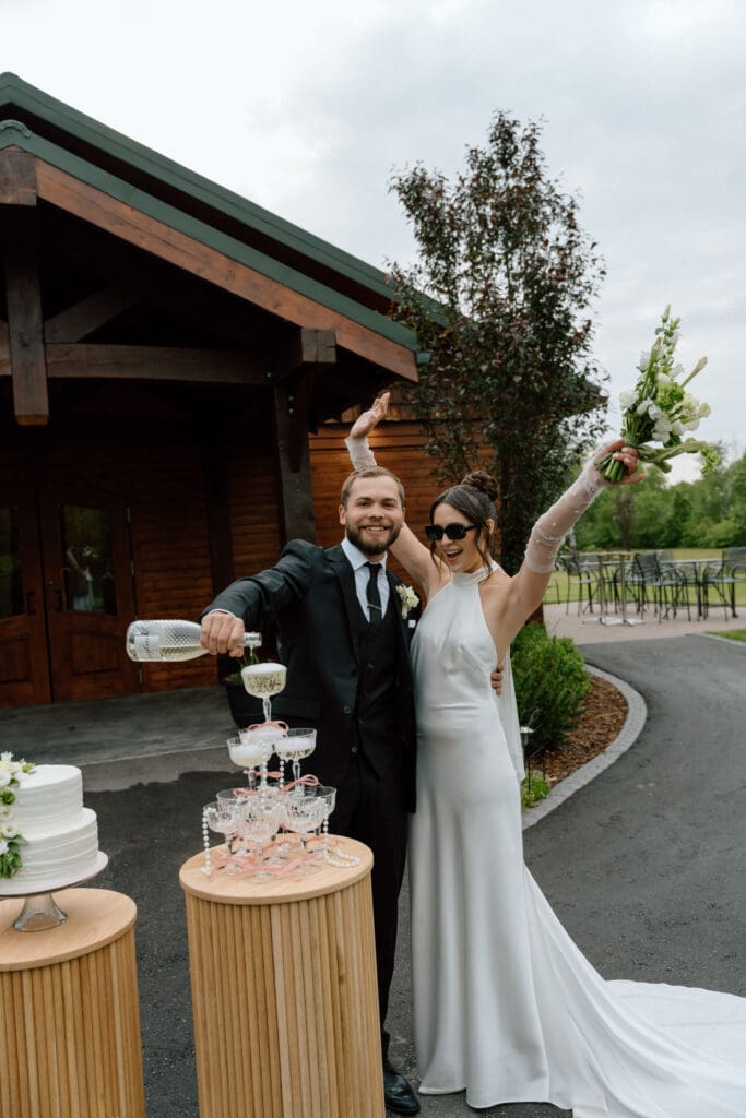 Groom pouring wine into glass tower while bride celebrates during their cottage core editorial wedding at Rivers Edge Resort Manitoba