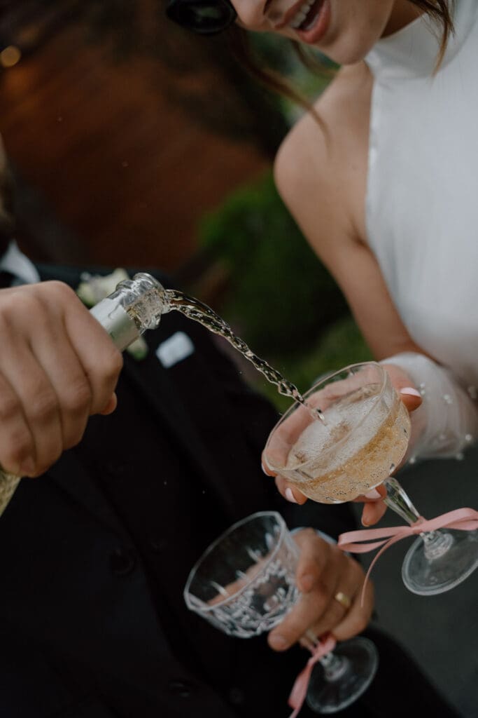 Close-up of groom pouring wine into bride's glass during intimate cottage core wedding reception at Rivers Edge Resort Manitoba