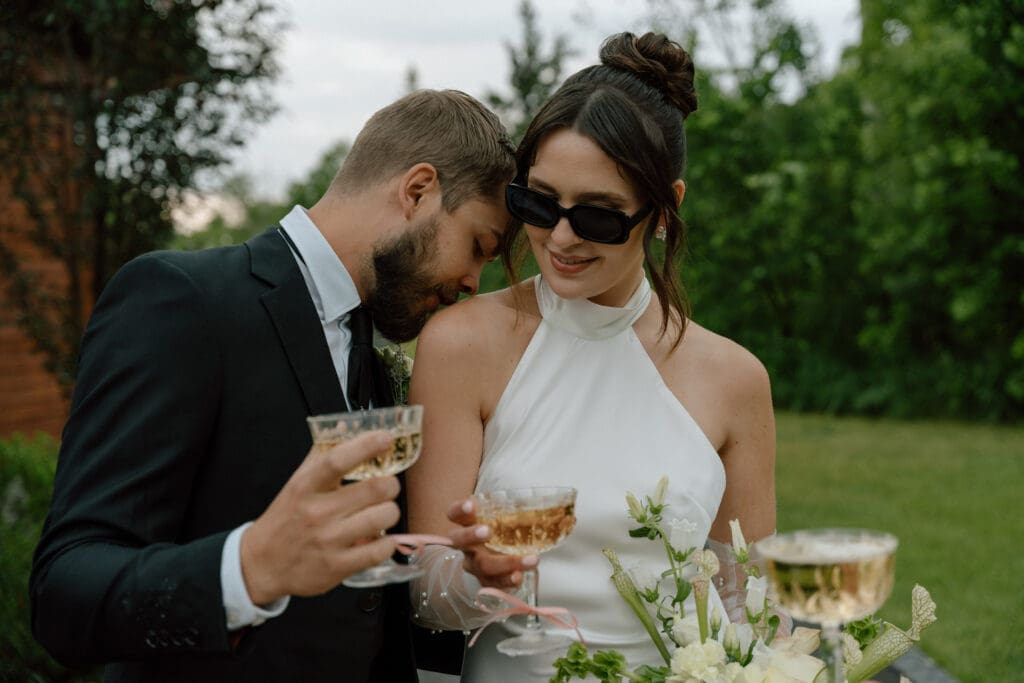 Romantic, candid moment of groom kissing bride's shoulder while the couple holds wine glasses at Rivers Edge Resort Manitoba