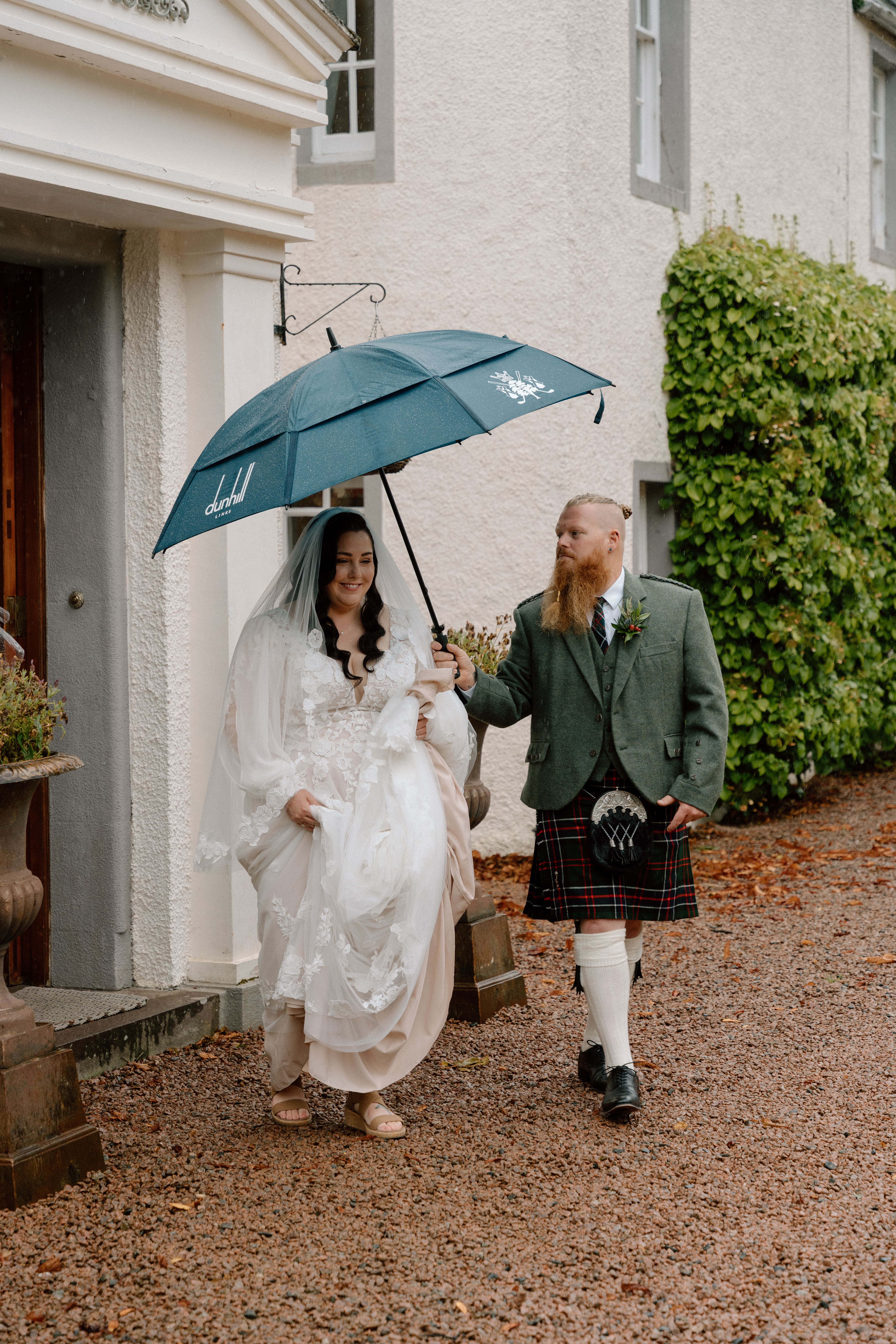 Bride being escorted by groom under a shared umbrella as they walk toward the wedding venue at Dunnottar Castle, Scotland.