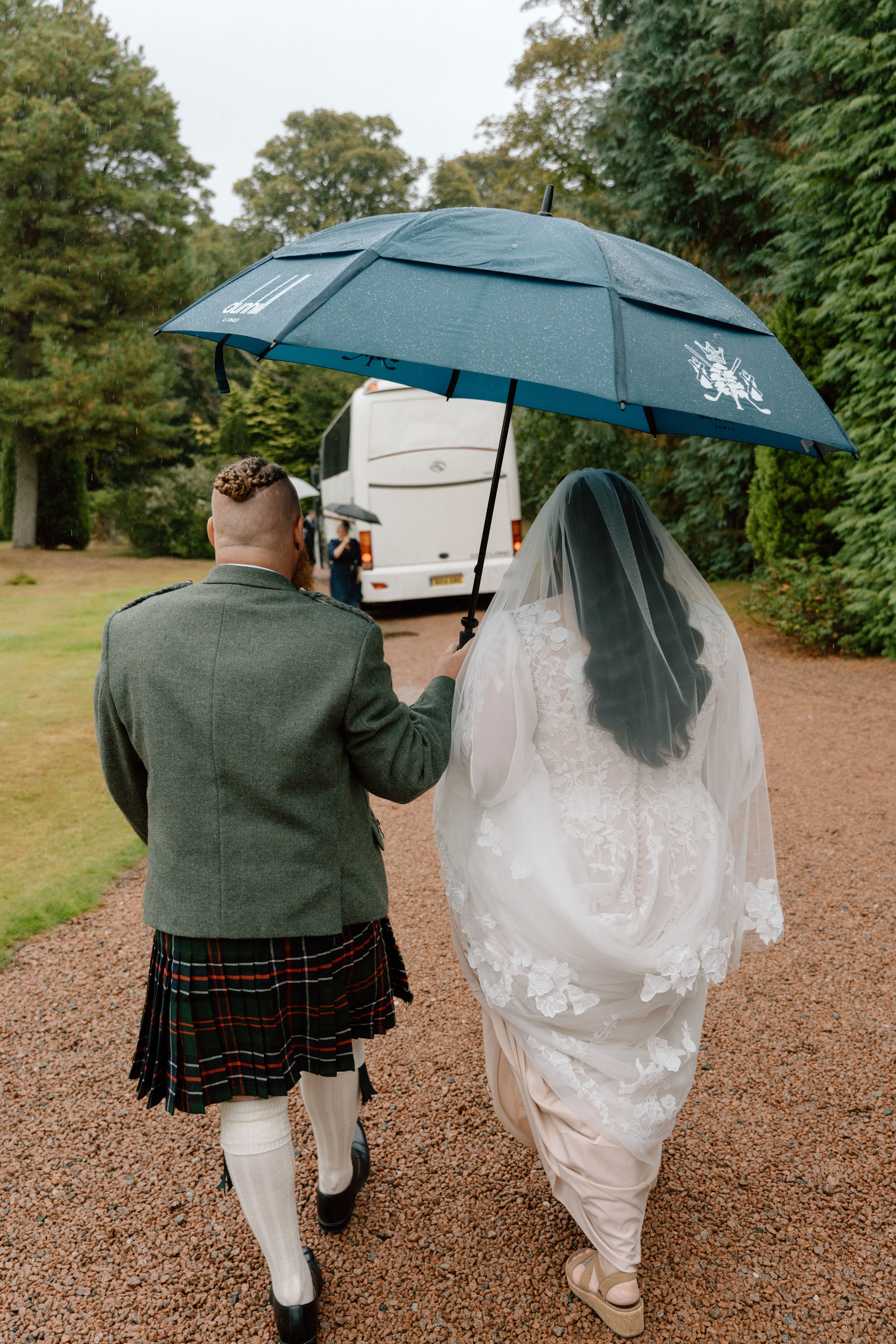 Groom gently guiding the bride with an umbrella as they walk to the bus for their wedding ceremony at Dunnottar Castle, Stonehaven.