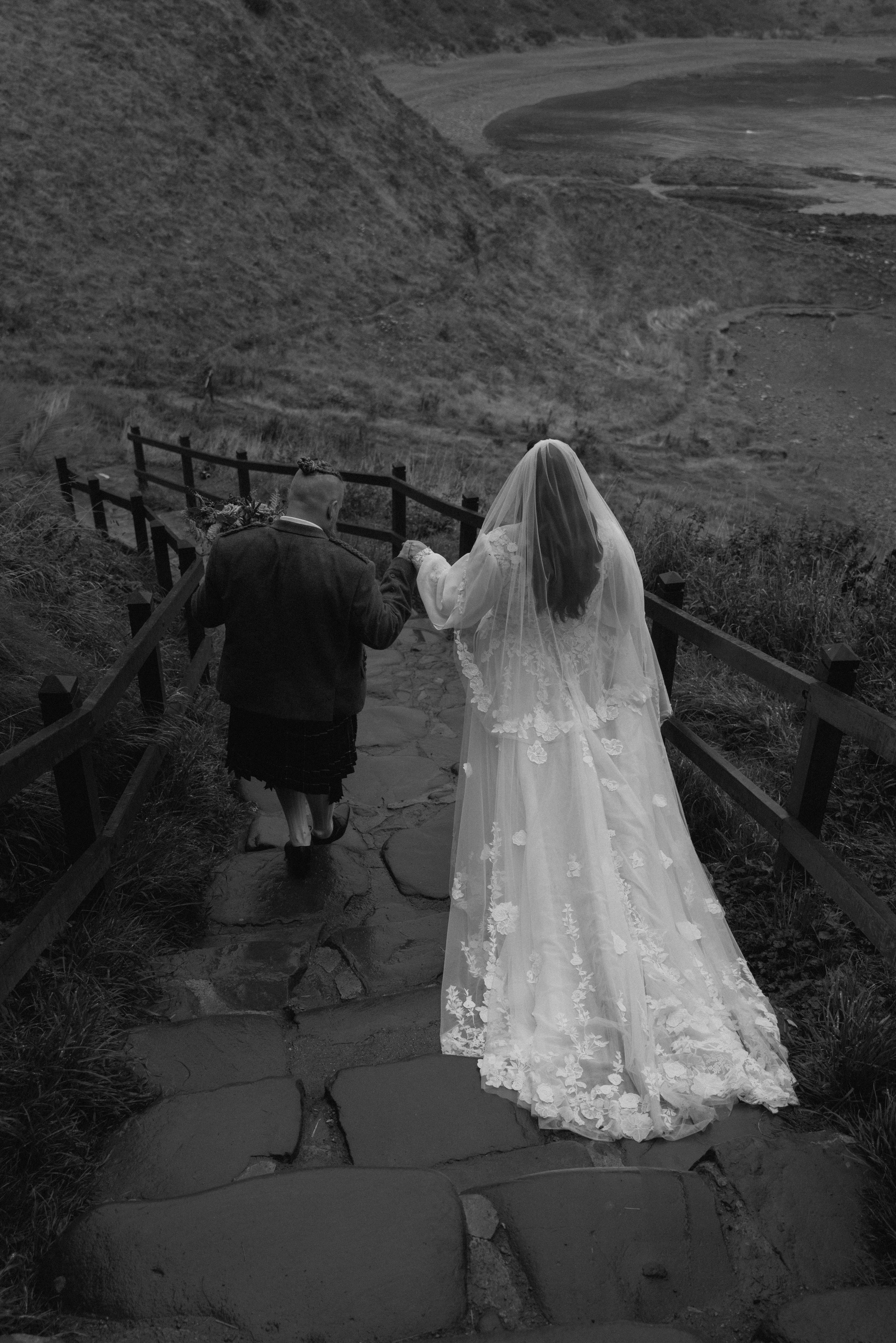 Groom escorting bride down stone steps near Dunnottar Castle with natural movement and candid emotion.