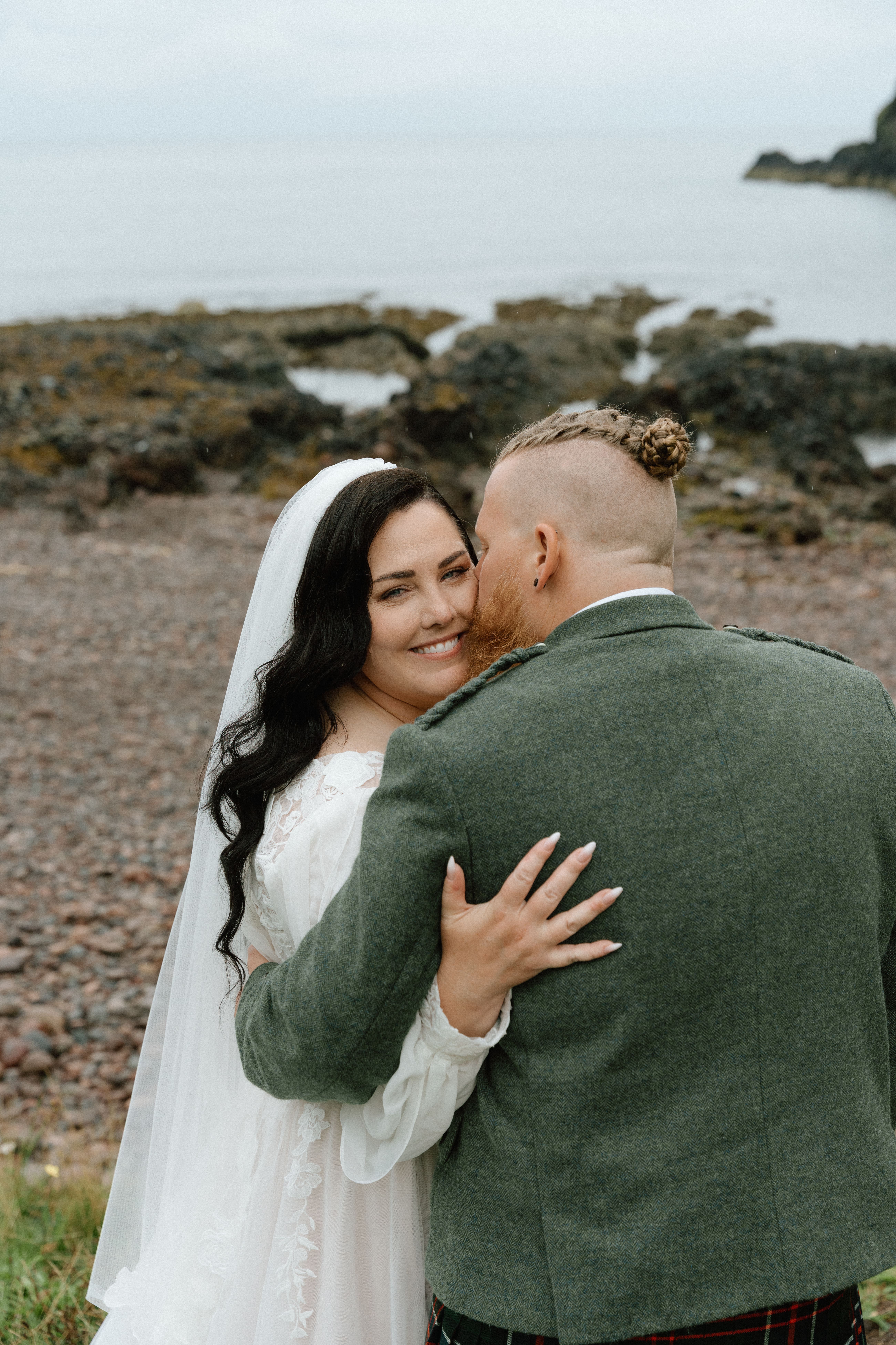 Candid wedding photography of the bride near Dunnottar Castle, Scotland, with the groom kissing the bride on the cheek at a rocky, coastal shoreline.
