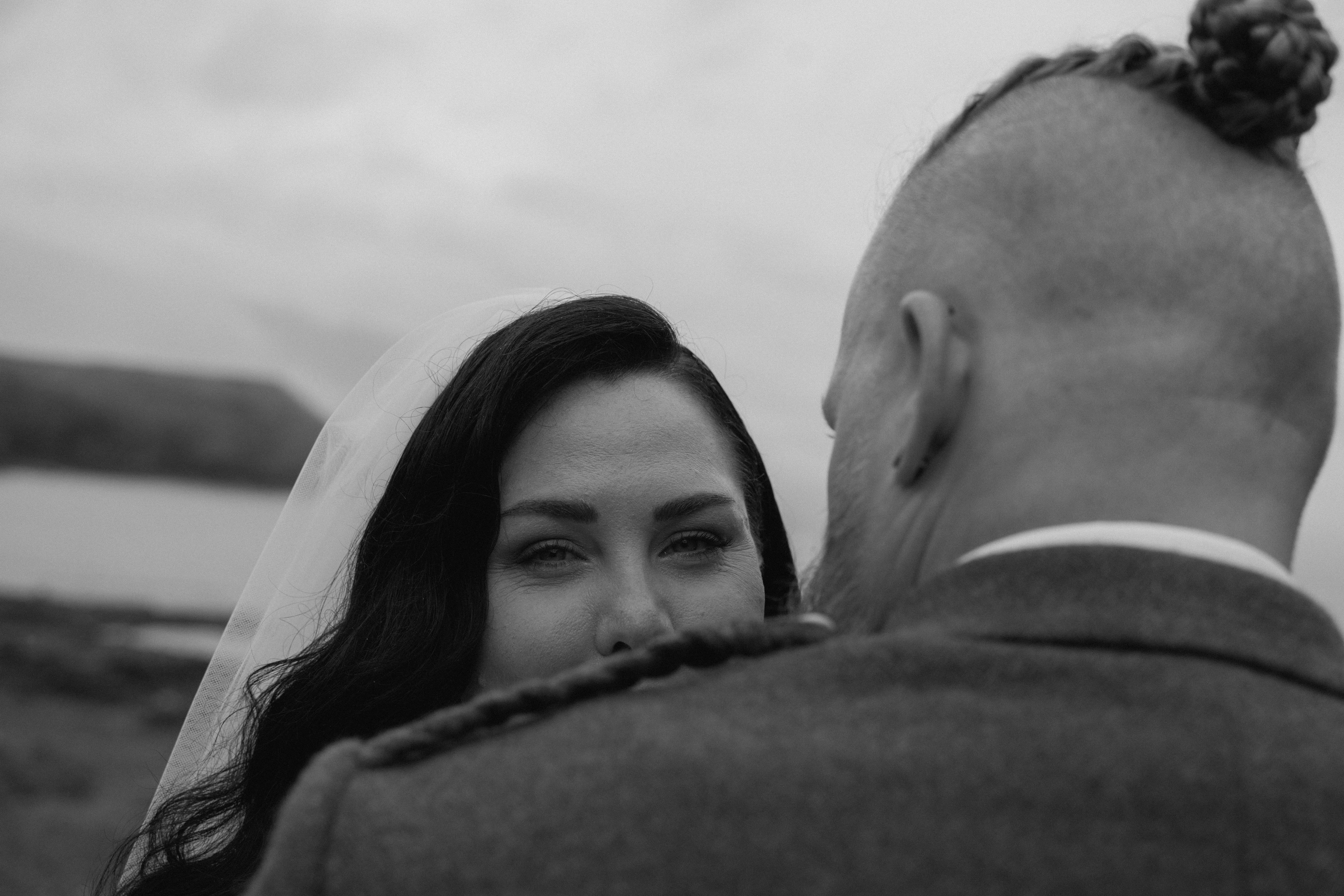 Documentary-style bridal portrait captured near Dunnottar Castle, showing the bride in focus while the couple shares a quiet, emotional moment in coastal shoreline.