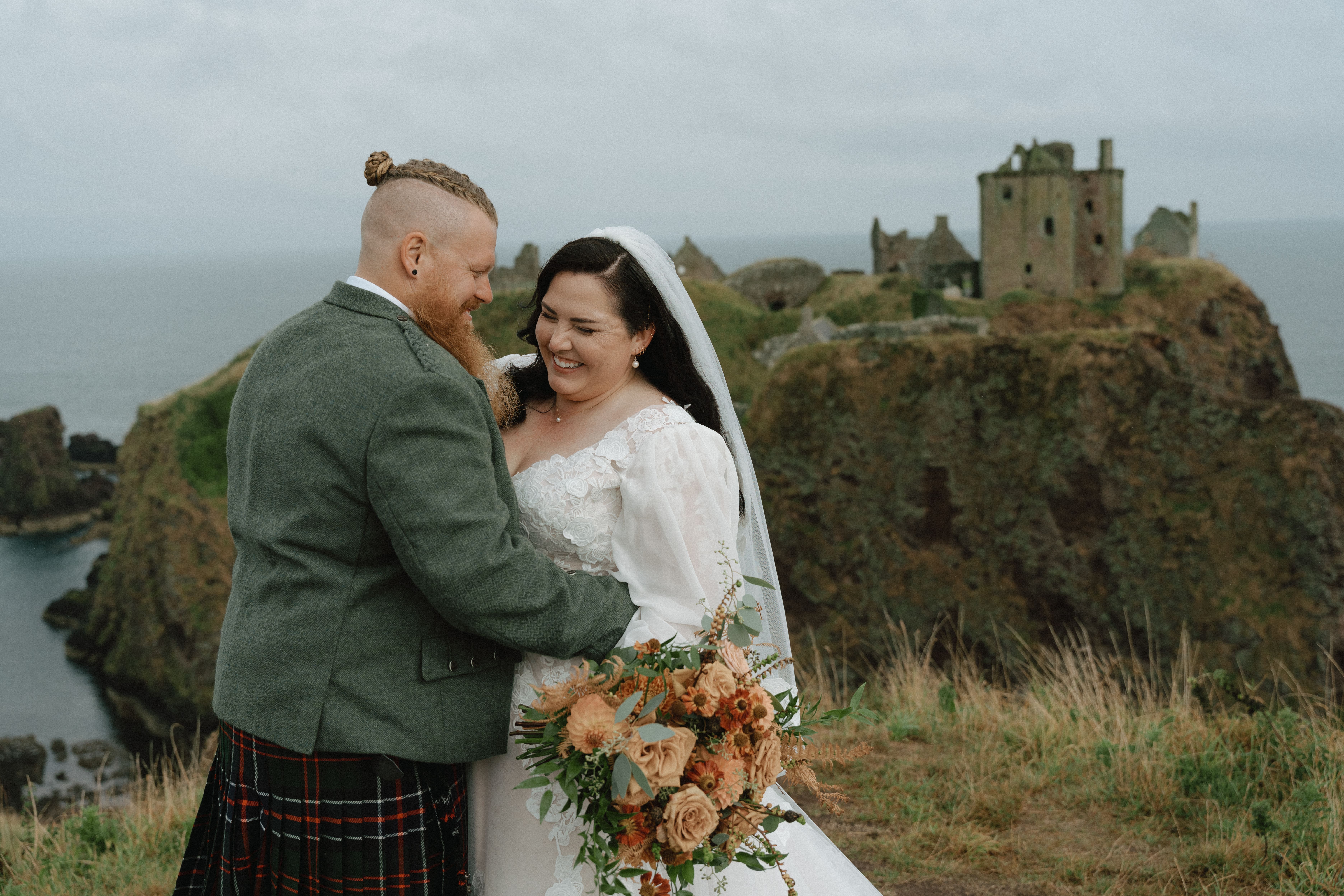 Natural documentary wedding portrait of a couple hugging while the bride laughs brightly on the coastal shoreline near Dunnottar Castle.