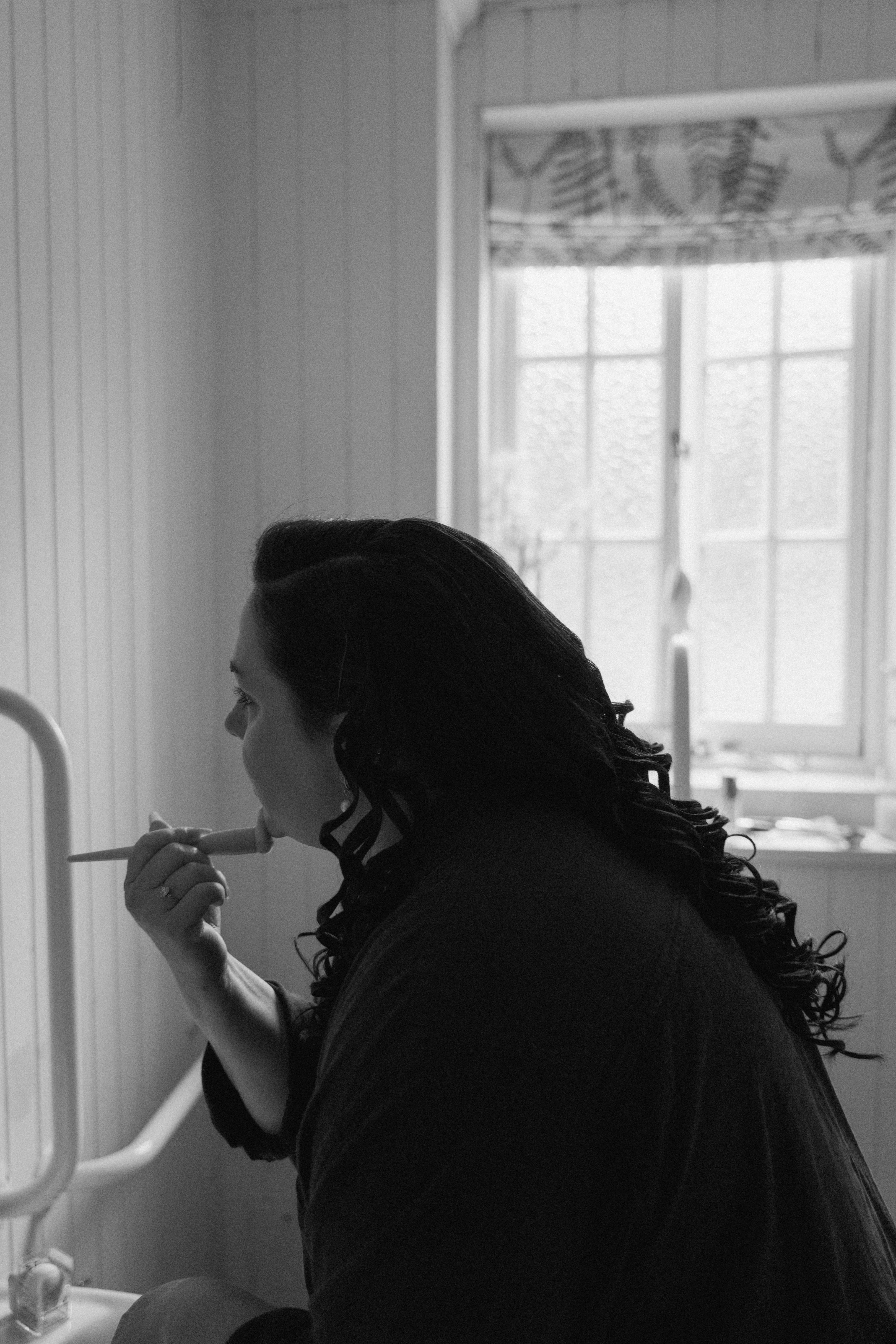 Documentary-style bridal preparation image of the bride doing her own makeup while looking into a mirror during a calm wedding morning in Scotland.