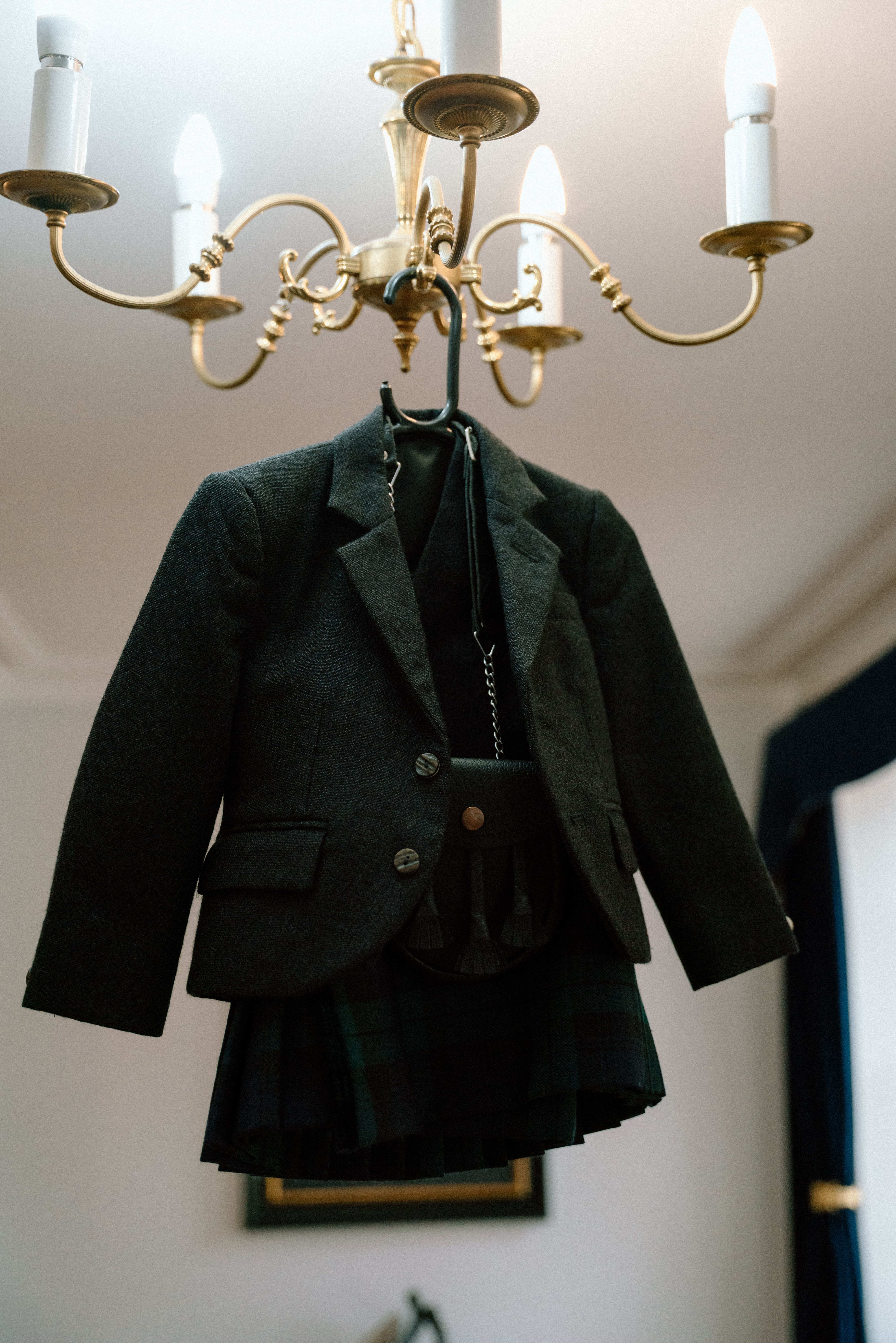 Fine art documentary wedding image of a tailored suit and kilt hanging elegantly from a chandelier, showcasing Scottish wedding details at Elsick House.
