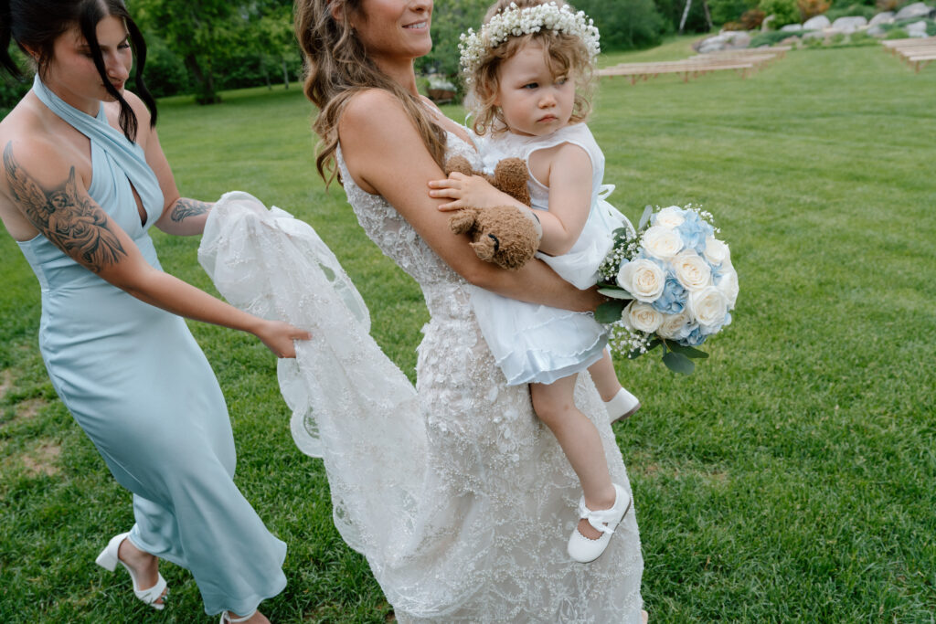Bride carrying a flower girl with a teddy bear and bouquet at an outdoor wedding