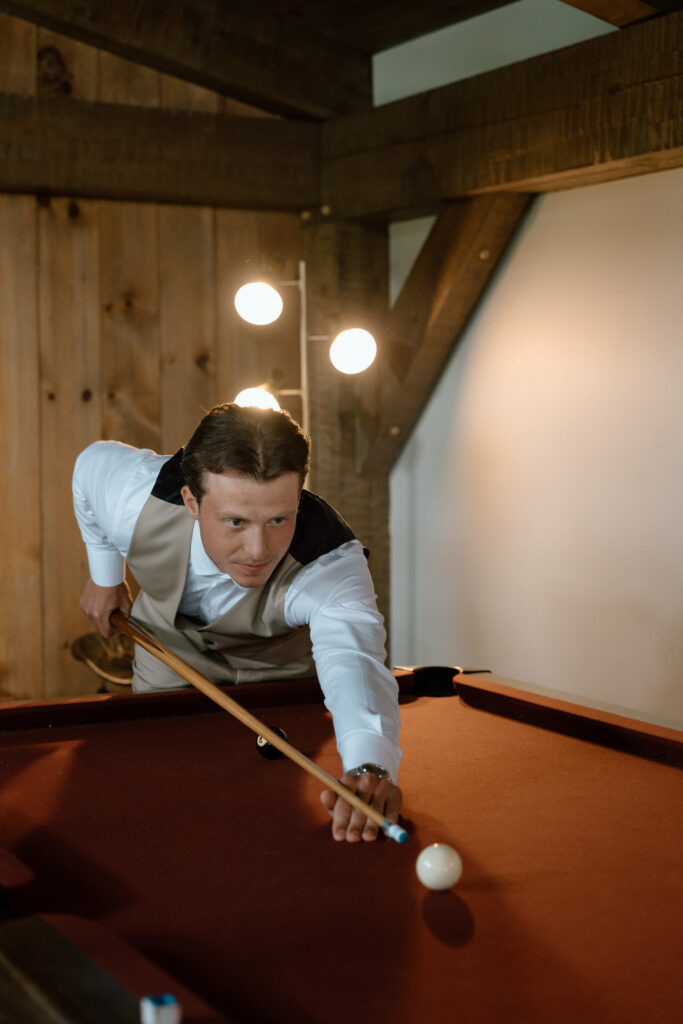 Groom in a white shirt and vest playing pool inside a rustic wedding venue at Hawthorne Estates, Manitoba