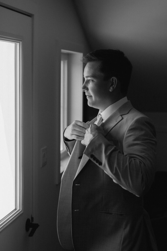 Groom adjusting his suit while looking out the window in a clean, editorial black and white wedding photograph