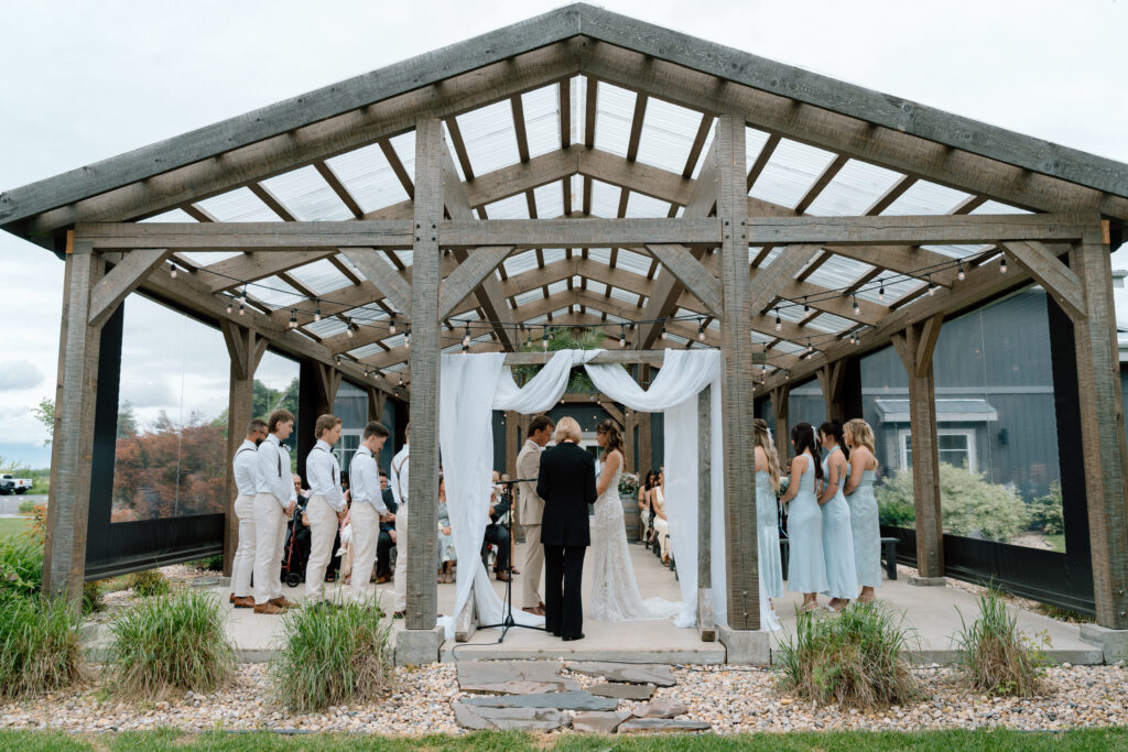 Bride and groom exchanging vows beneath a draped wedding arch at an outdoor barn ceremony with their wedding party
