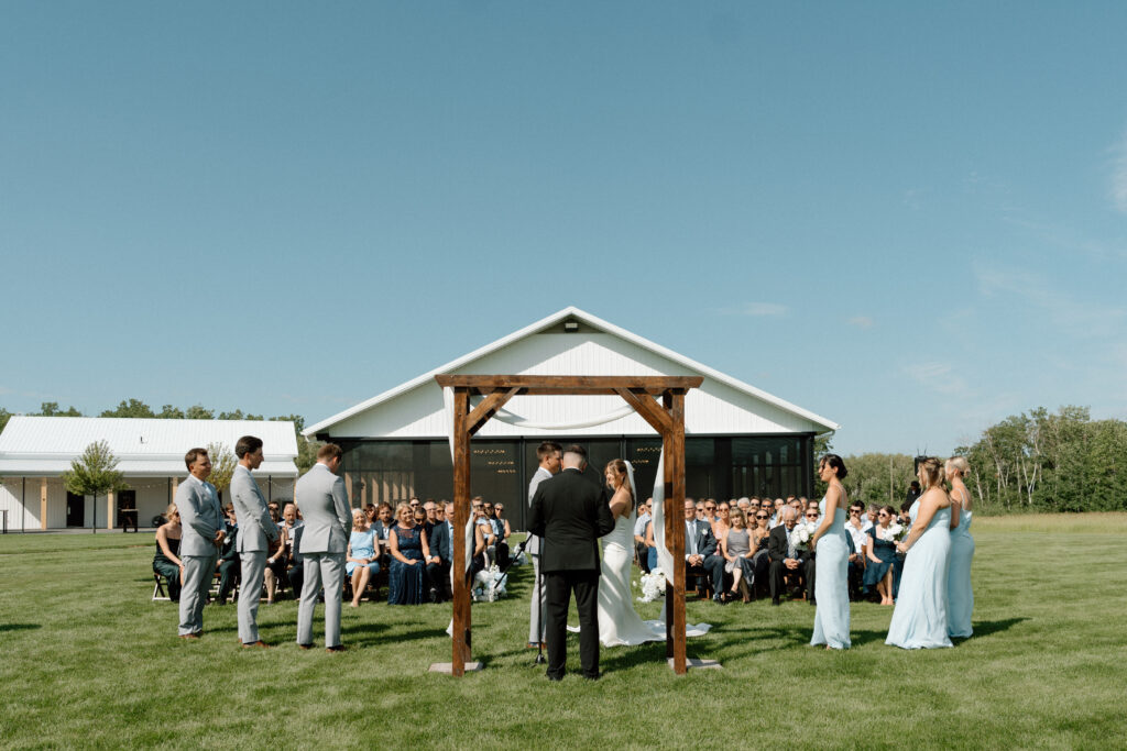 Wide view of Julianna and Cole's outdoor wedding ceremony at The White Poplar beneath a bright blue Manitoba sky